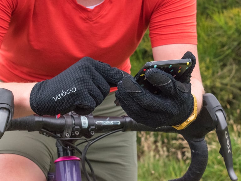 Black Velocio TRAIL Gloves holding a phone over a gravel bike's handlebar, logo on the back of the hand.