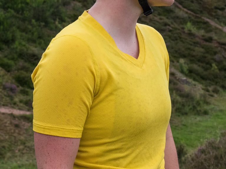 Close-up of the yellow Velocio Radiator TRAIL Tee's V-neck and perforated texture, with faint mud marks.