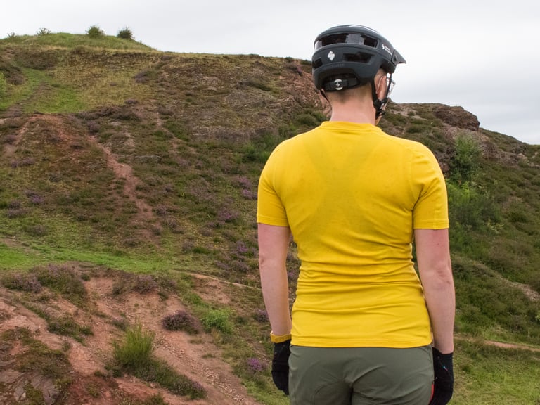 Rear view of the yellow Velocio Radiator TRAIL Tee worn with a trail helmet on a hillside.