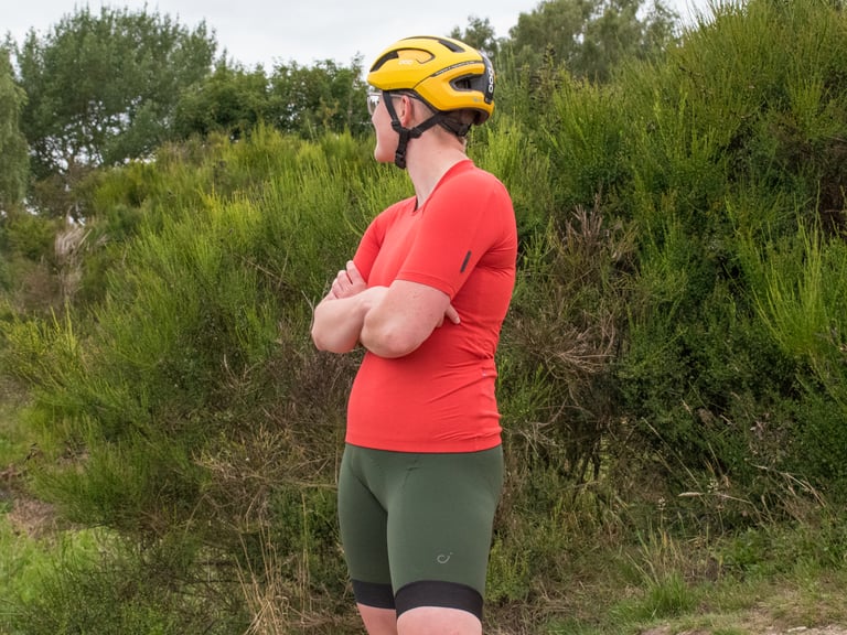 Rider in Velocio LUXE khaki bib shorts, red jersey and yellow helmet, standing with arms folded beside green scrub.