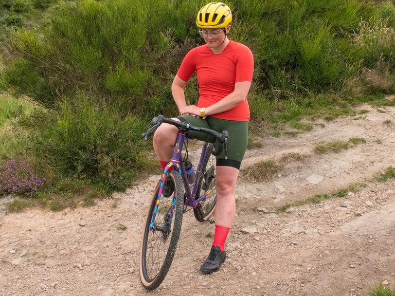Rider in the Velocio Micromodal Jersey and green bib shorts stands over a purple gravel bike on a stony track.