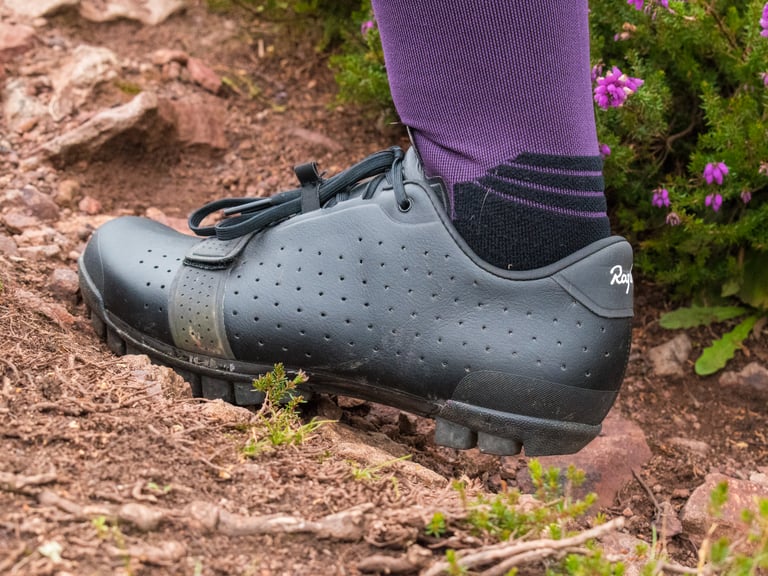 Rider walking in the Rapha Explore Shoes, with heel and sole visible, on a rocky heather-lined trail.