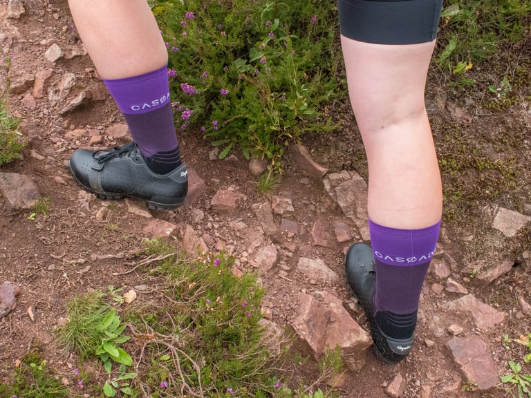 Rear view of purple two-tone Cascada Cross Socks with white logo at the ankle, worn with Rapha shoes on a rocky path.