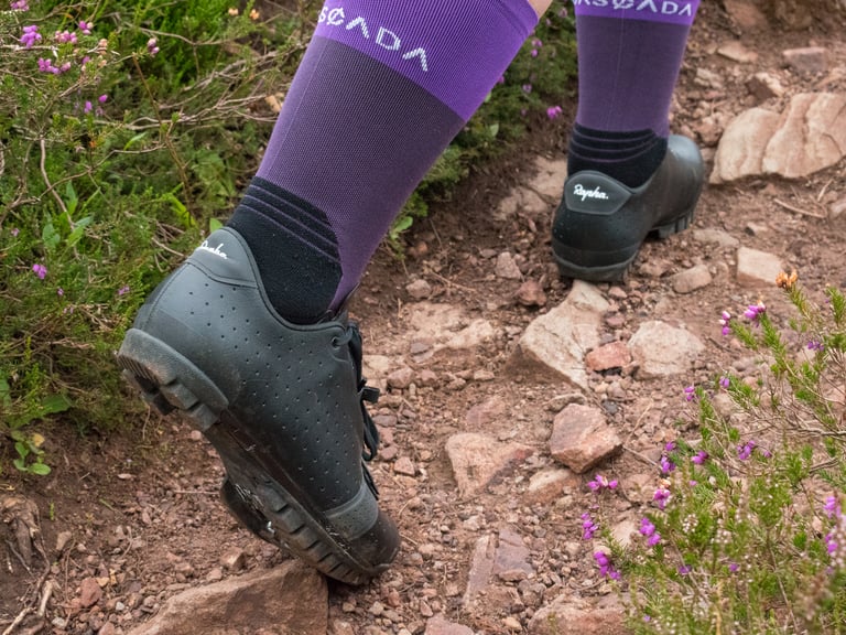 Rider in Rapha Explore Shoes and purple socks steps up a rocky heather trail, seen from behind.