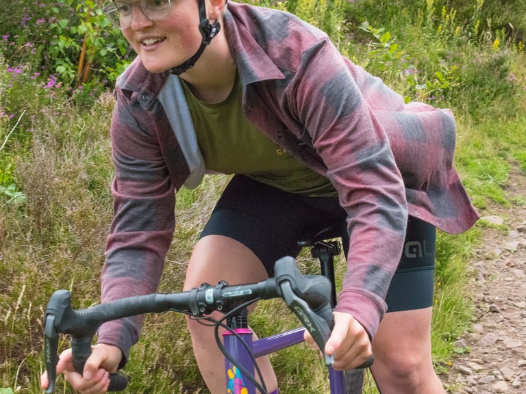 Rider on a purple gravel bike wears the Cascada Land Wool Shirt over a green Trail Tee on a grassy hillside track.