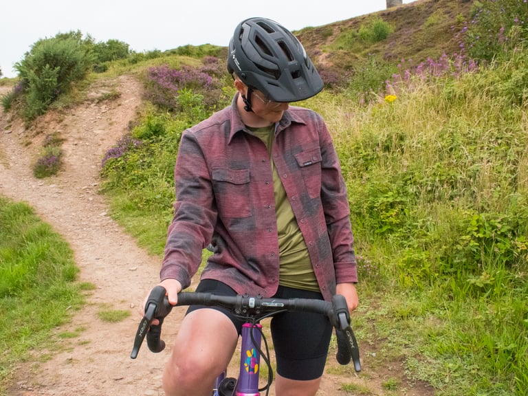 Rider in the burgundy and grey check Cascada Land Wool Shirt pauses on a gravel bike on a hillside path among heather.