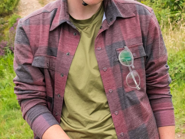 Clear-framed glasses hooked into the left chest pocket of the Cascada Land Wool Shirt, worn over a green tee.