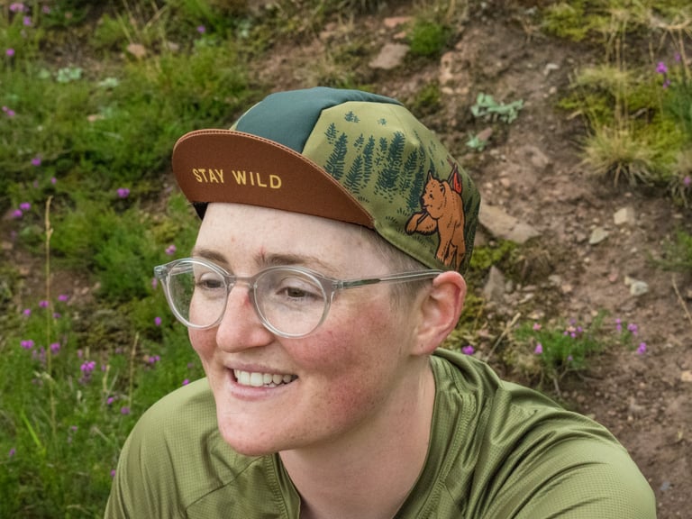 Smiling rider wears the Cascada Stay Wild cycling cap, with a bear illustration on the side panel and brown peak.