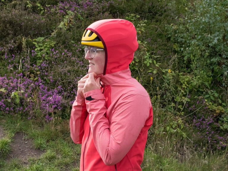 Rider cinches the stretchy hood of the Infinium Hybrid Jacket over a yellow helmet beside purple heather.