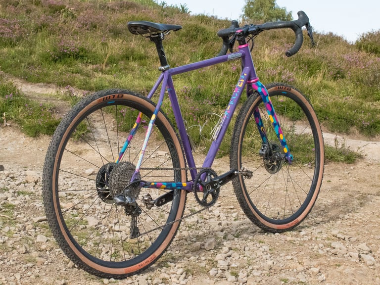 Purple custom steel Mercredi cyclo-cross bike with tan-wall WTB tyres, parked on a stony track beside heather.