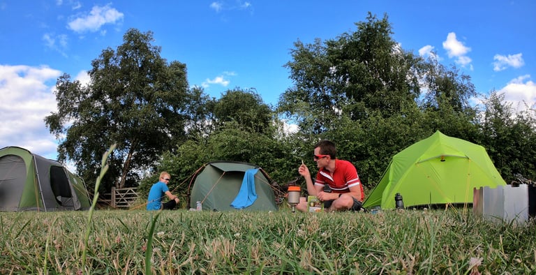 Two riders sit cooking beside three tents on the grass at the Hickling campsite under a blue summer sky.