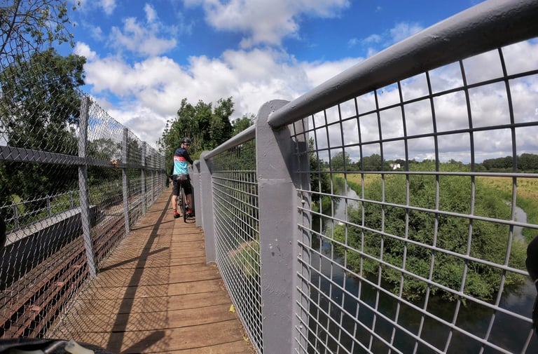 Rider pauses on a mesh-sided bridge over a Broads river, with the narrow gauge Bure Valley Railway tracks alongside.