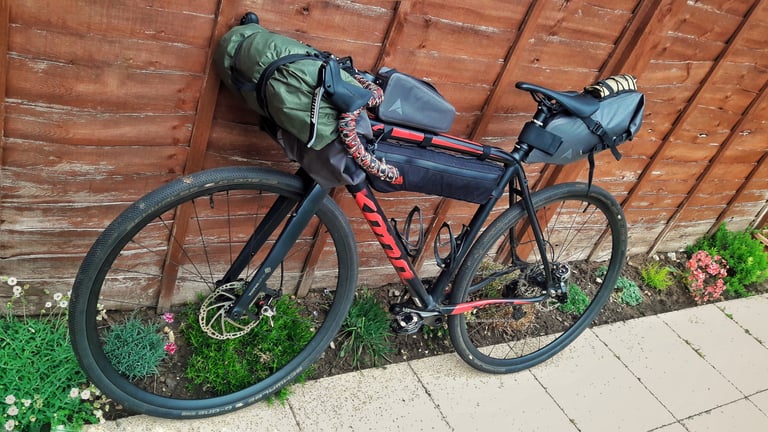 Kona Private Jake gravel bike fully loaded with Alpkit frame bag, bar roll and saddle pack, leaning against a fence.
