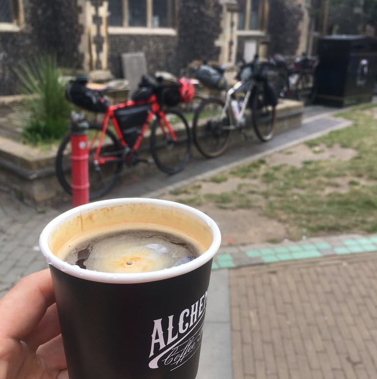 Black coffee in a paper cup in Norwich, with loaded bikepacking bikes parked against a low wall behind.