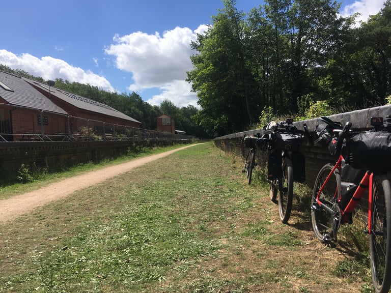 Three loaded bikes lean on a brick wall beside the gravel path of Marriott's Way, with brick sheds across the trail.