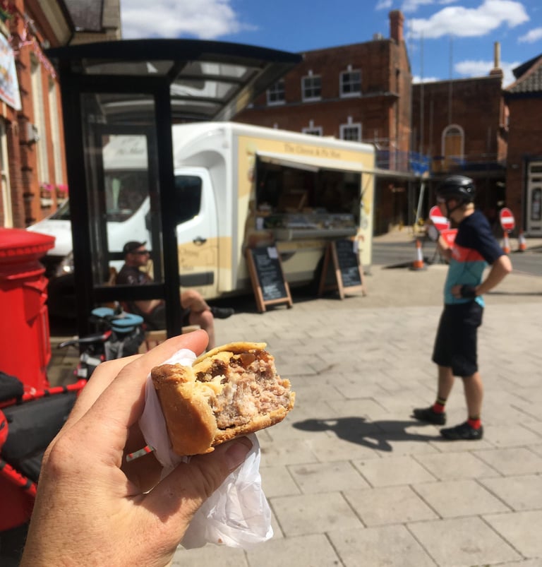 Half-eaten pork pie in hand in Aylsham, with the Cheese and Pie Man van and a cyclist behind in the square.