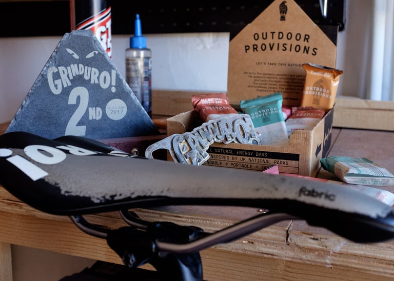 Outdoor Provisions energy bars, a Grinduro slate trophy and bottle opener on a workbench behind a saddle.