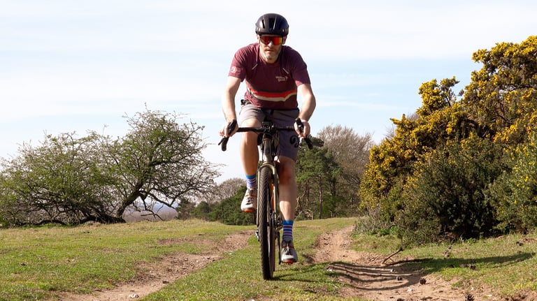 Rider on a gravel bike wearing the Stolen Goat Merino Gravel Jersey on a grassy track beside flowering gorse.