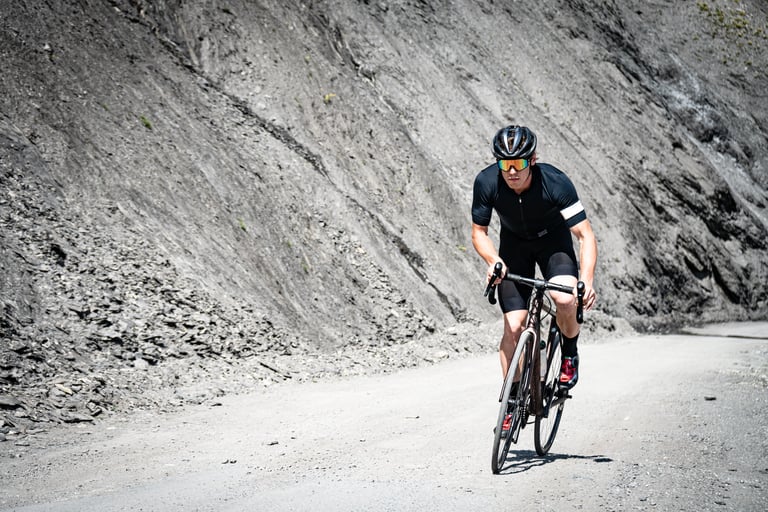 Rider wearing SunGod Vulcans sunglasses climbs a gravel road beneath a grey rock face.