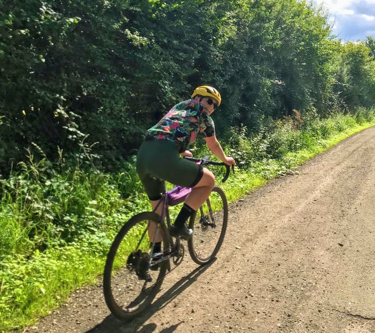 Katherine rides a purple gravel bike along a dry gravel track in Velocio LUXE bib shorts, looking back over her shoulder.