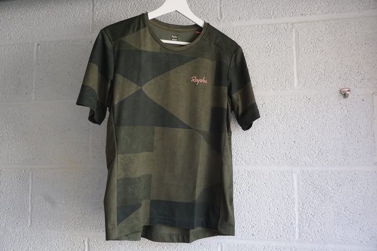 Rapha women's Technical T-Shirt in a green digi-camo print on a hanger against a white breeze-block wall.