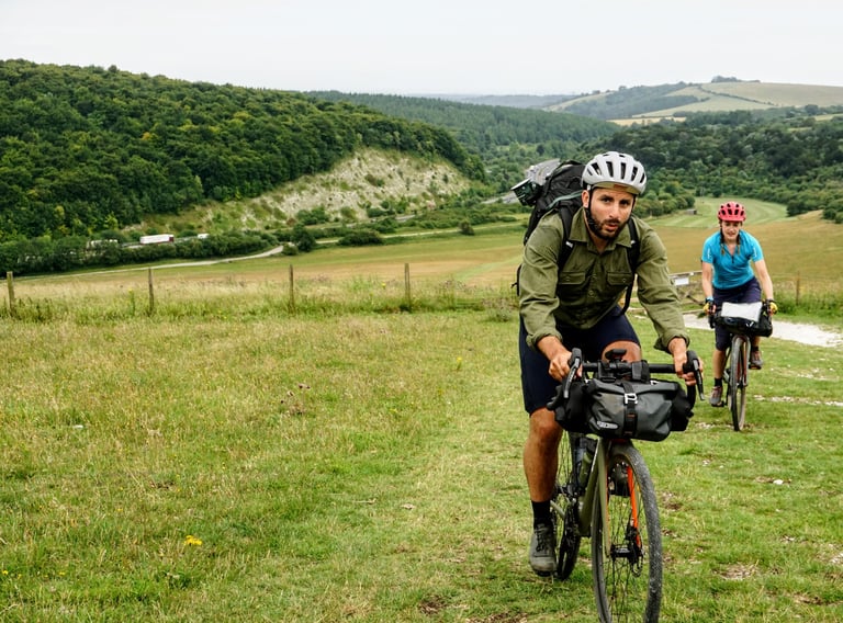Two riders on loaded gravel bikes climb Butser Hill on the South Downs Way, with the main road below.