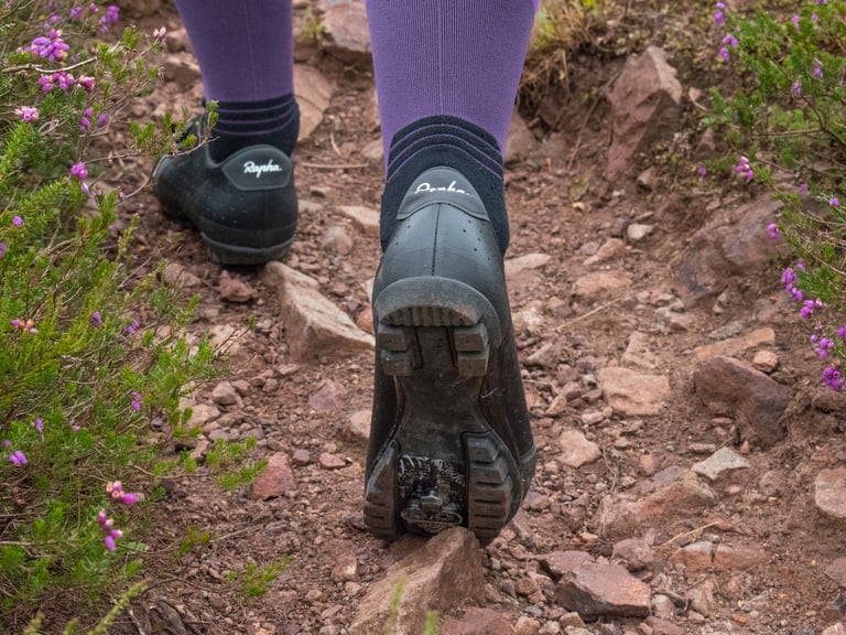 Rear view of a rider in Rapha Explore Shoes walking a rocky trail, showing the rubber tread and cleat recess.