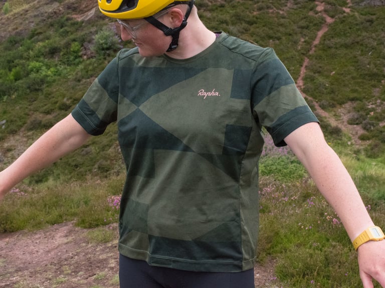 Reviewer wears the loose-fitting Rapha Technical T-Shirt in forest green print with a yellow helmet on a heather hillside.