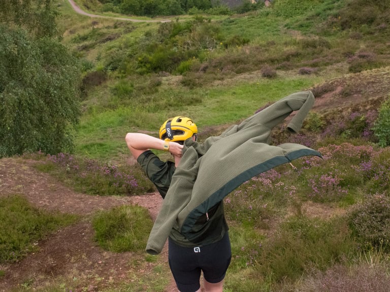 Rider in a yellow helmet throws on the olive Rapha Explore Technical Hoodie on a heathery hillside trail.