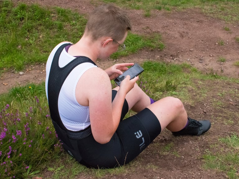 Katherine sitting on a grassy track in Alé Stones cargo bib shorts, typing notes on a phone mid-test.