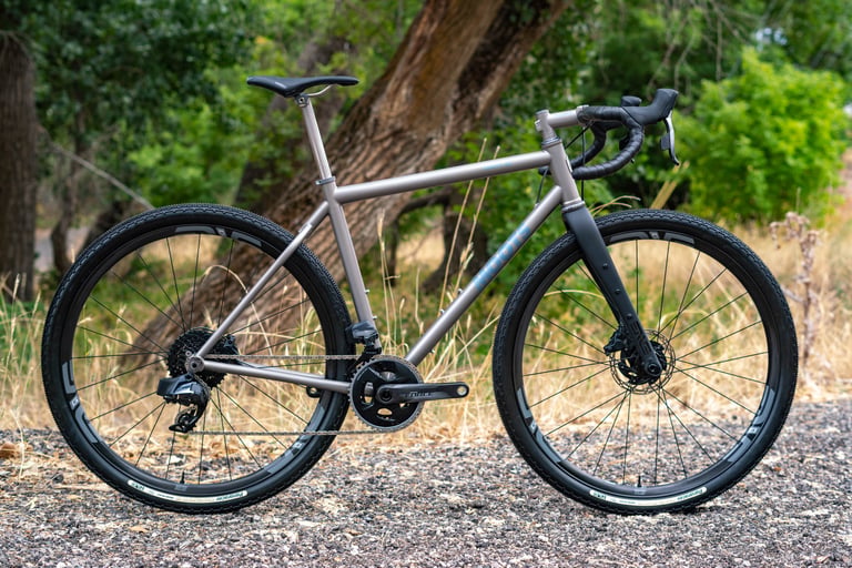 Moots titanium gravel bike with a black ENVE Adventure Fork and SRAM Force drivetrain, parked on gravel among trees.