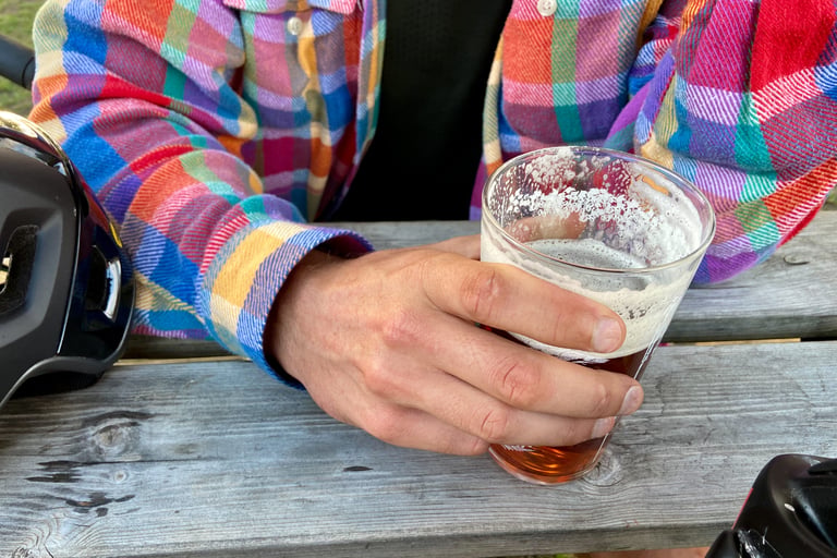 Hand in a bright flannel shirt holding a pint on a wooden table beside a helmet, the post-ride drinks square.