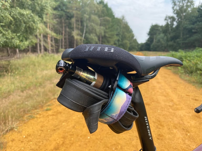 Granite PUNK inflator, tool roll and inner tube strapped under a saddle on a sandy forest track.