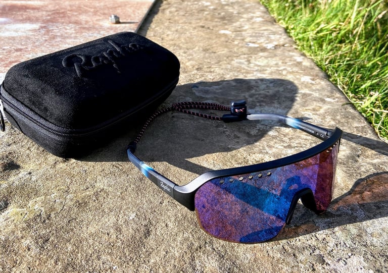 Rapha Explore Glasses with a purple-blue mirrored lens and neck strap on a stone slab beside their black zipped case.