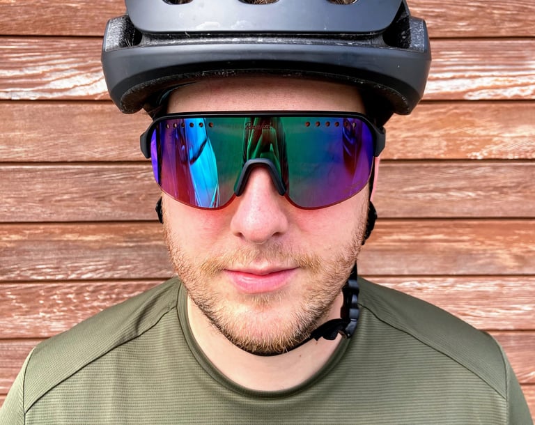 Rider faces the camera in Rapha Explore Glasses, the mirrored purple-green lens with a row of vent holes along its top edge.
