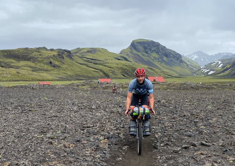 Bikepacker rides over a black lava field away from the red-roofed Alftavant huts, another rider following behind.