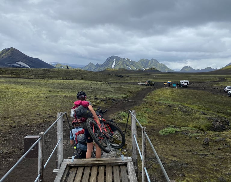 Bikepacker shoulders a bike over a wooden footbridge, a companion behind, under grey skies on the Iceland Divide.