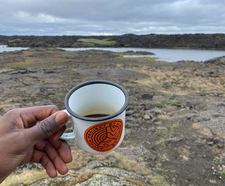 Hand holds an enamel mug of coffee over a lava field and lake on the last day of the Iceland Divide.