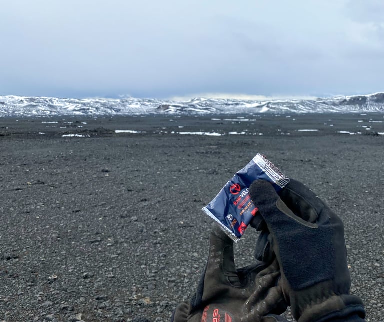 Gloved hand holds a Rawvelo chocolate bar above a black gravel plain with snowy mountains on the Iceland Divide.