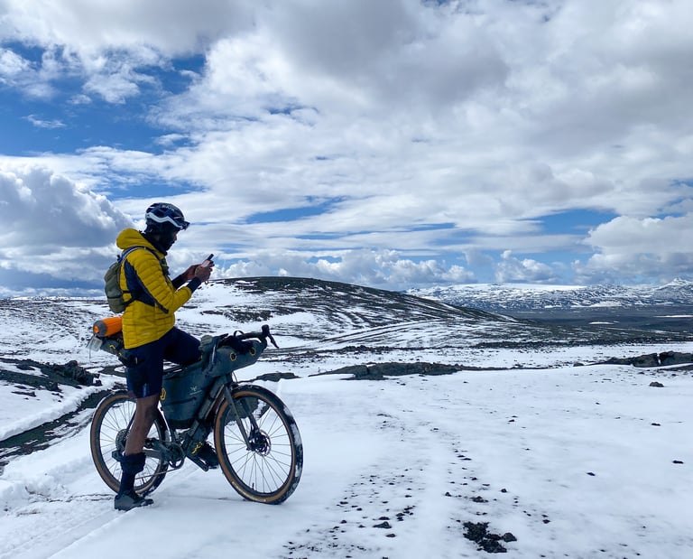 Bikepacker in a yellow down jacket checks a phone beside a loaded bike on a snowy track in Iceland.