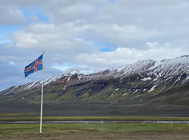 Icelandic flag flies in front of snow-streaked mountains and a dark lava plain on the Iceland Divide.