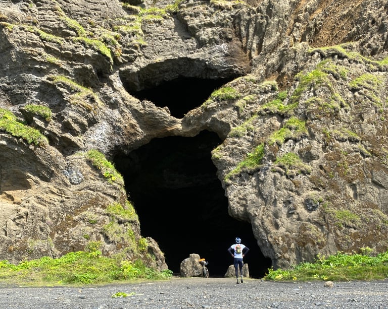 Rider and bike dwarfed by the mouth of Yoda Cave, a huge dark opening in a mossy rock face in south Iceland.