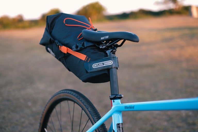 Ortlieb seat pack with orange bungee cords strapped under a WTB saddle on a light blue gravel bike in low sun.