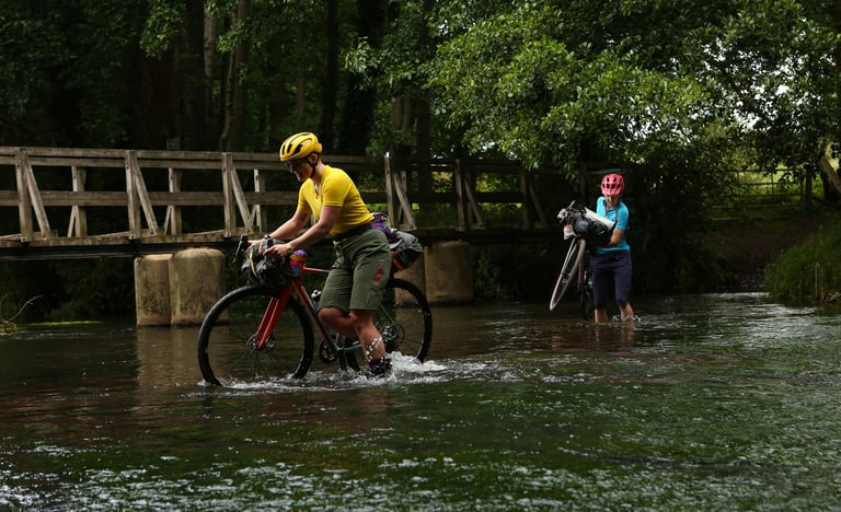 Two bikepackers wade the River Wey near Farnham on the King Alfred's Way, one carrying a loaded bike across.