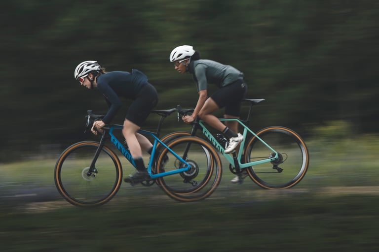 Two women ride Canyon gravel bikes at speed against a blurred forest, previewing the women's King's Cup gravel race.