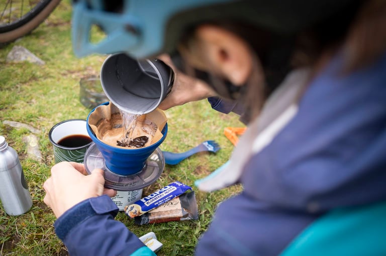 Rider pours hot water through a coffee dripper on the grass beside a mug and snack bar, brewing up on the trail.