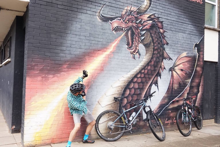 Rider in a check shirt shields their face from a fire-breathing dragon mural, two bikes leaning on the wall, South Wales.