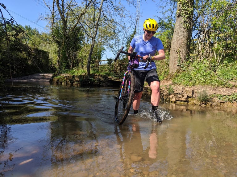 Smiling rider in a yellow helmet carries a gravel bike through a shallow ford near Bristol.