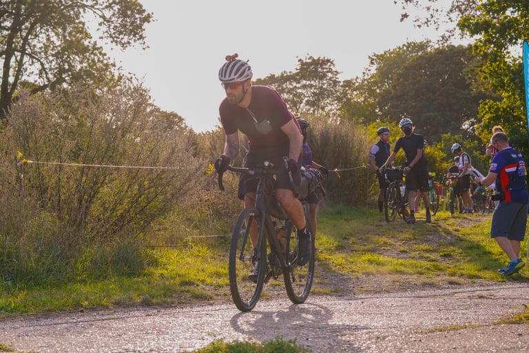 Rider in a maroon jersey leaves the staggered start of the Great British Escapade as marshals and riders look on.