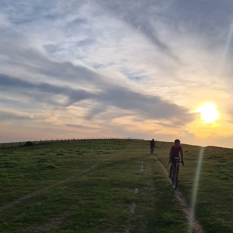 Two Great British Escapade riders cross a grassy ridge towards a low sun under streaked clouds.
